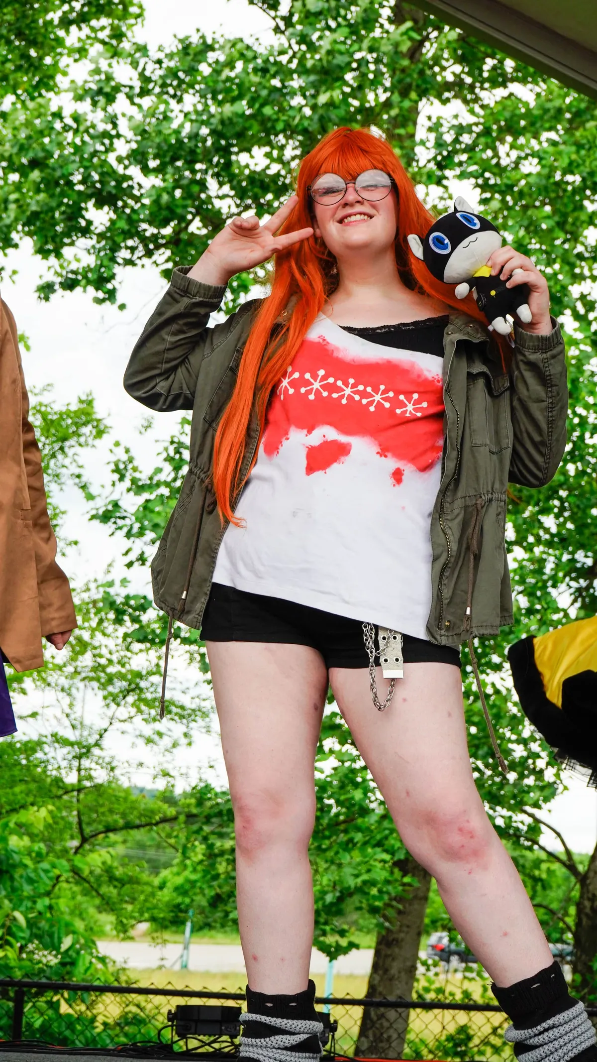 A cosplayer with a long orange wig, round glasses and a green jacket flashes a peace sign while holding a black cat plush on an outdoor stage at Knox Pop Con.
